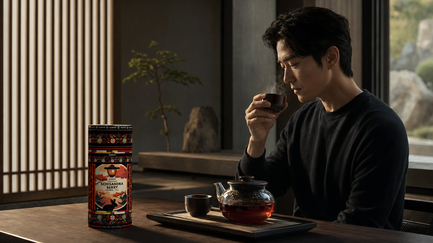 Man enjoying tea beside KORYO Schisandra Berry Herbal Tea packaging and glass teaware in a refined minimalist interior.