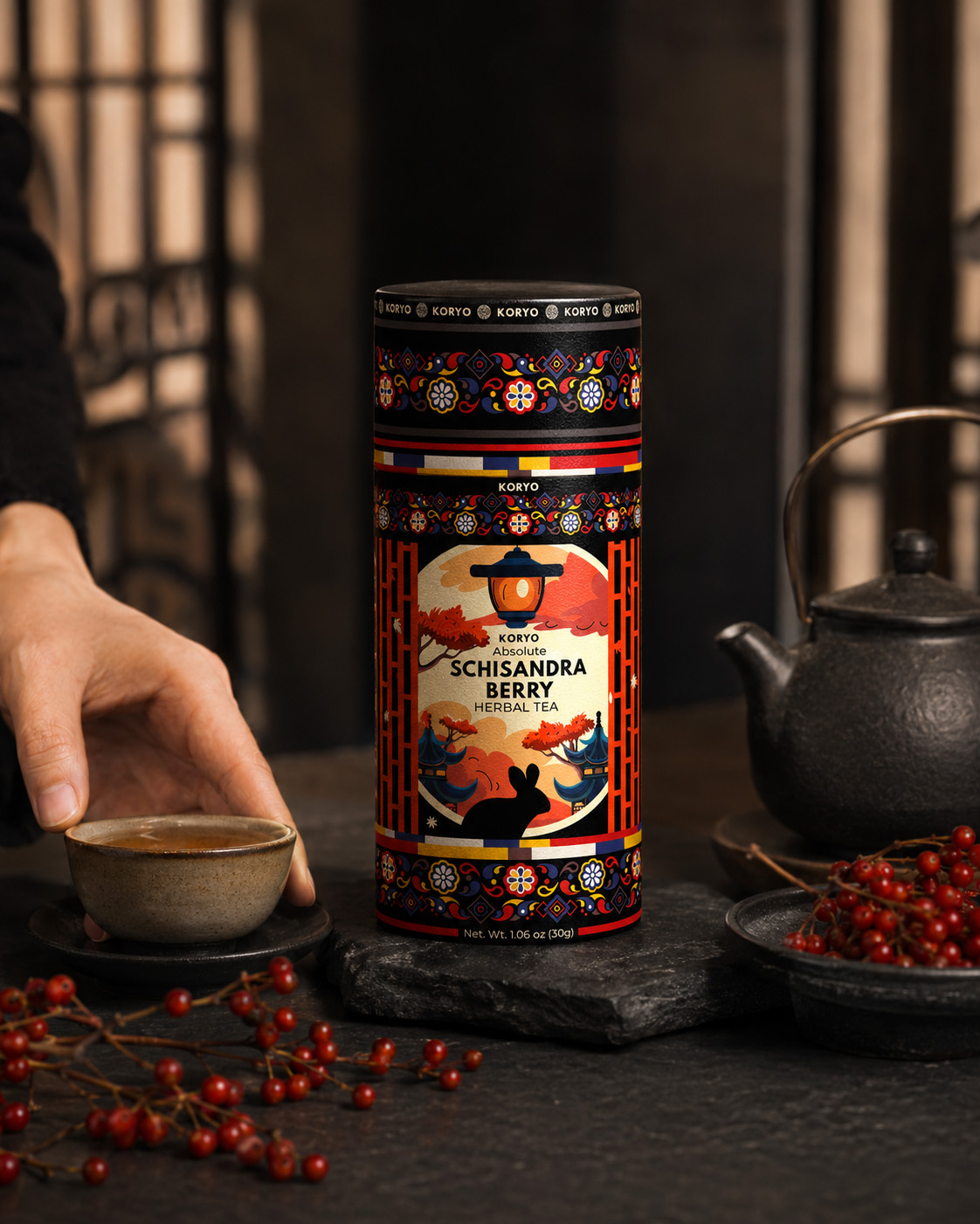KORYO Schisandra Berry Herbal Tea canister with a hand placing a tea cup beside dark teaware and red Schisandra berries.