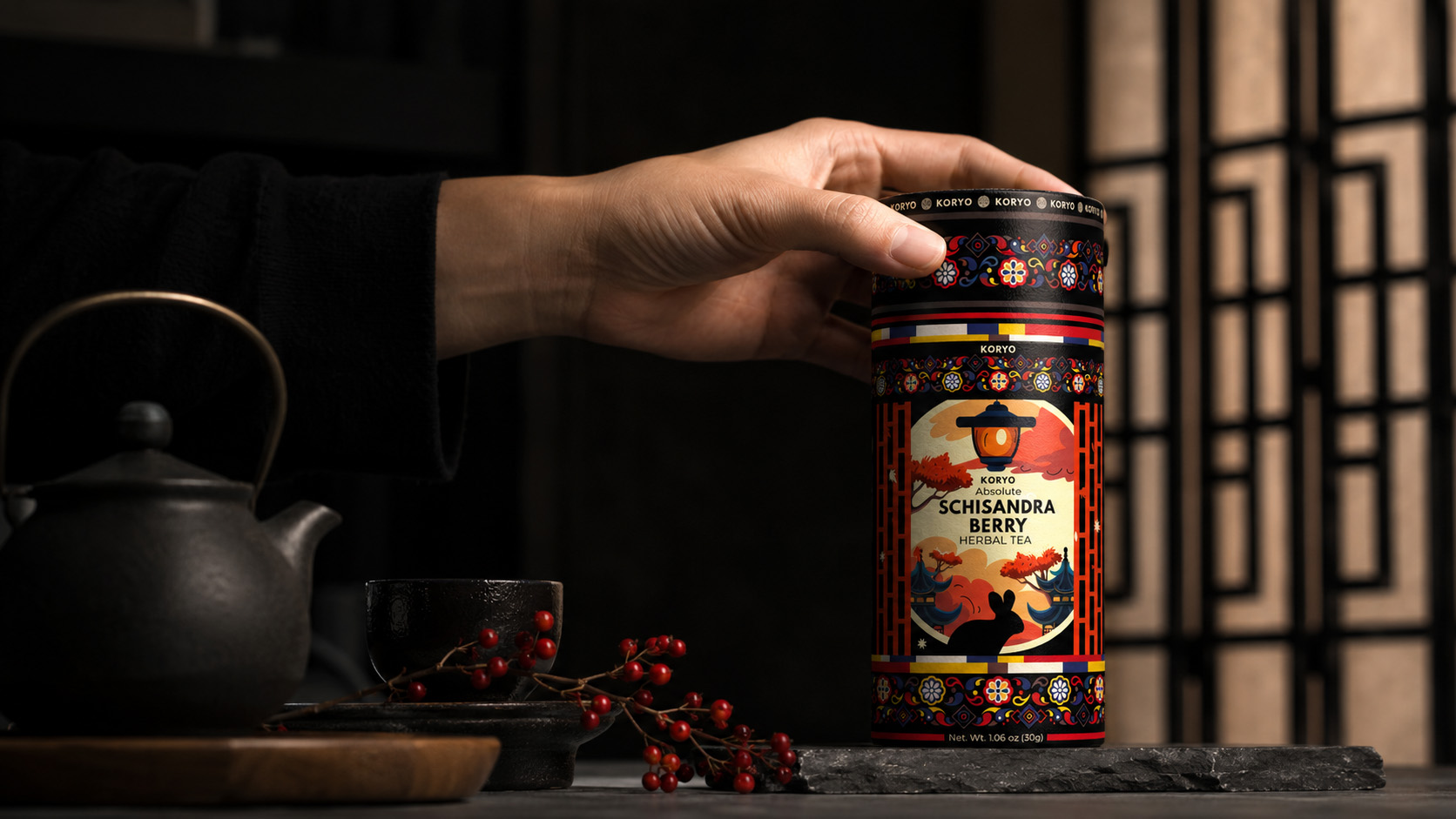 Hand reaching for KORYO Schisandra Berry Herbal Tea packaging beside dark teaware and red Schisandra berries in a warm Korean-inspired interior.