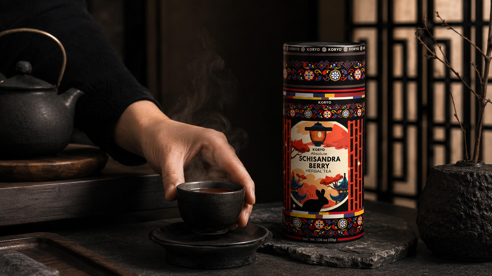 Hand reaching for a steaming cup beside KORYO Schisandra Berry Herbal Tea packaging and dark teaware in a warm Korean-inspired setting.