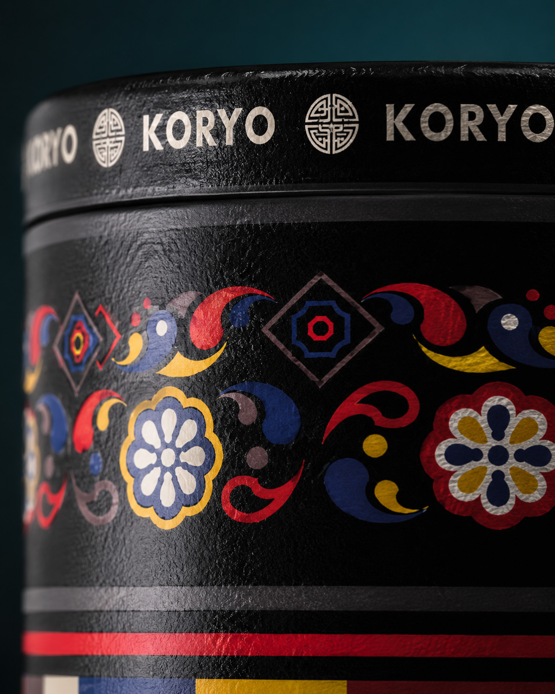 Macro photograph of the KORYO tea canister showing the textured lid rim, repeating wordmark, brand symbols, and Korean-inspired ornamental pattern.