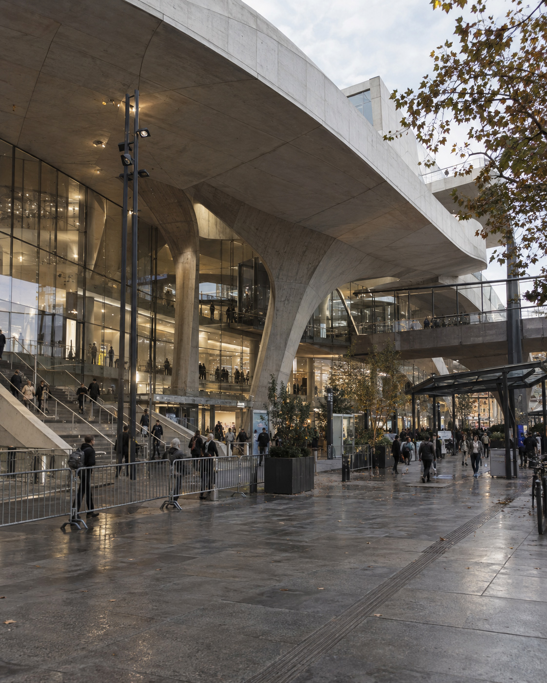 Contemporary public architecture with exposed concrete, glazed façades, layered circulation, stairs, sheltered walkways, and people occupying the space during wet overcast conditions.
